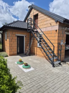 realizacja-kraty-schody-dom-jednorodzinny-3 - Loch - Kraty pomostowe Wema Producent Brick house with a black metal exterior staircase leading to a second-floor door and a paved courtyard below.